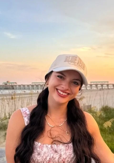 Young woman smiling at the beach during sunset, wearing a white baseball cap, a floral dress, and layered necklaces, with a pier in the background.