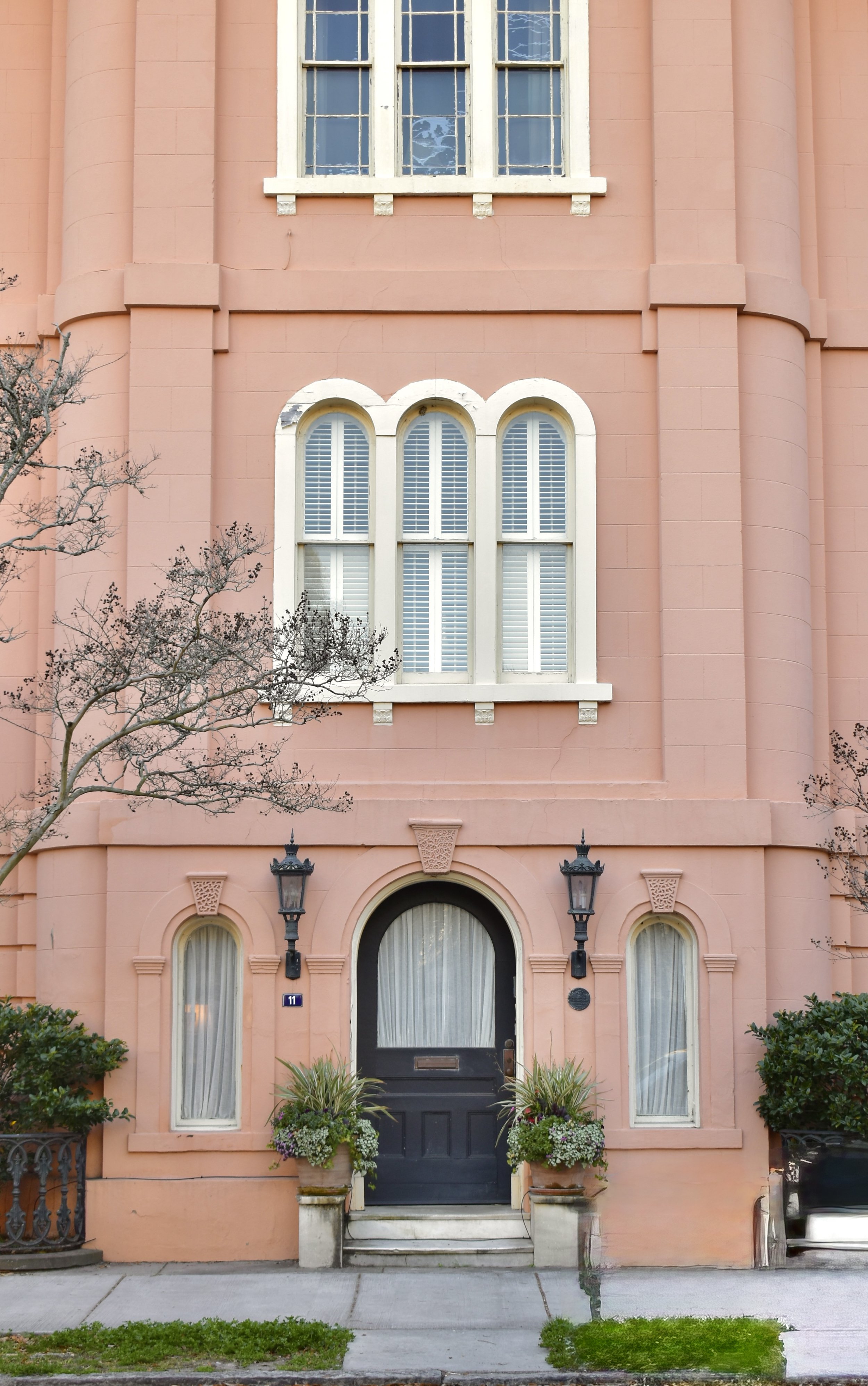 Pink building with arched windows, black door, and outdoor lanterns, with some trees and plants in front.
