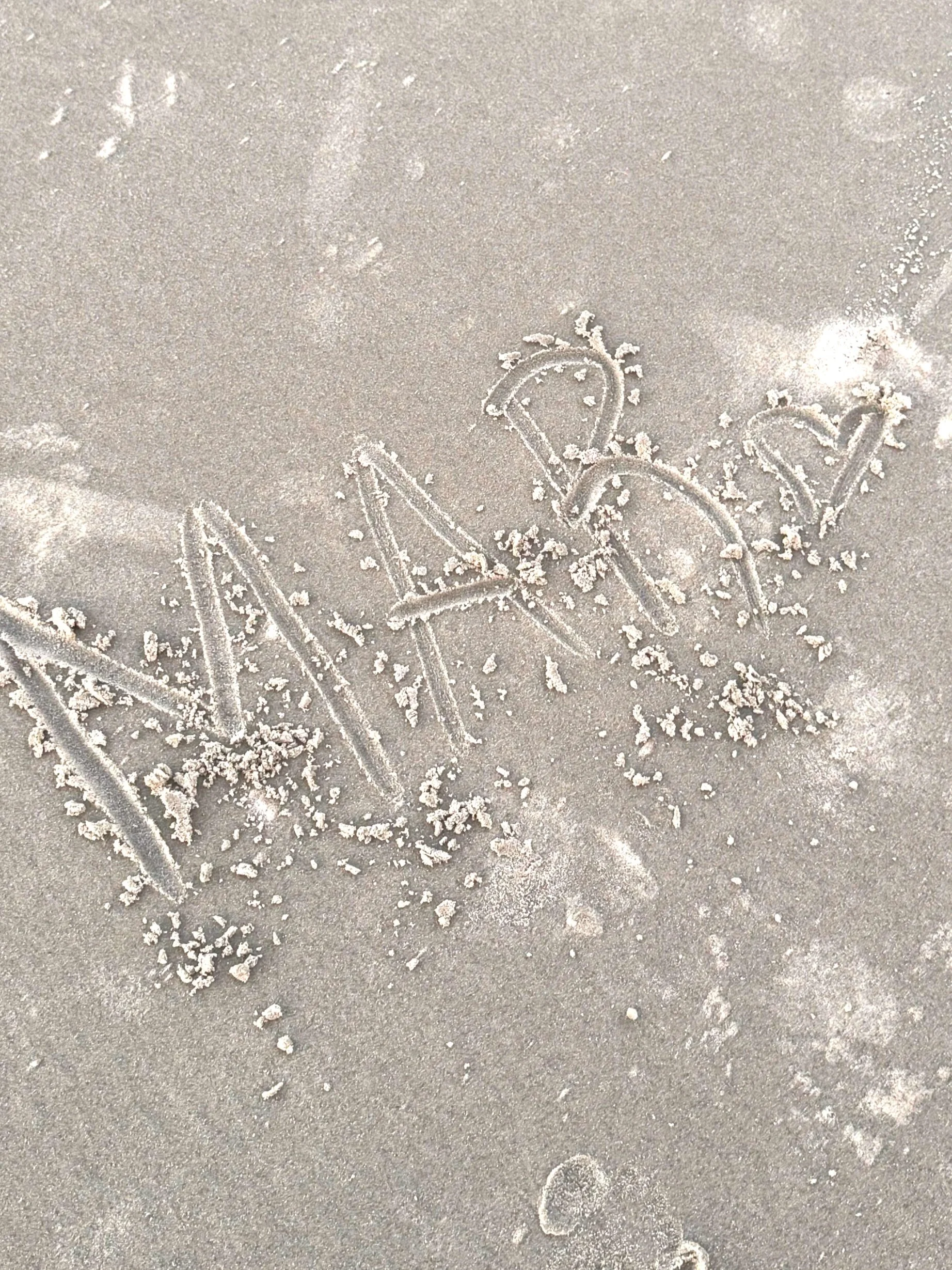 The word 'NAKED' is written in the sand with a stick, with some small grains of sand scattered around.