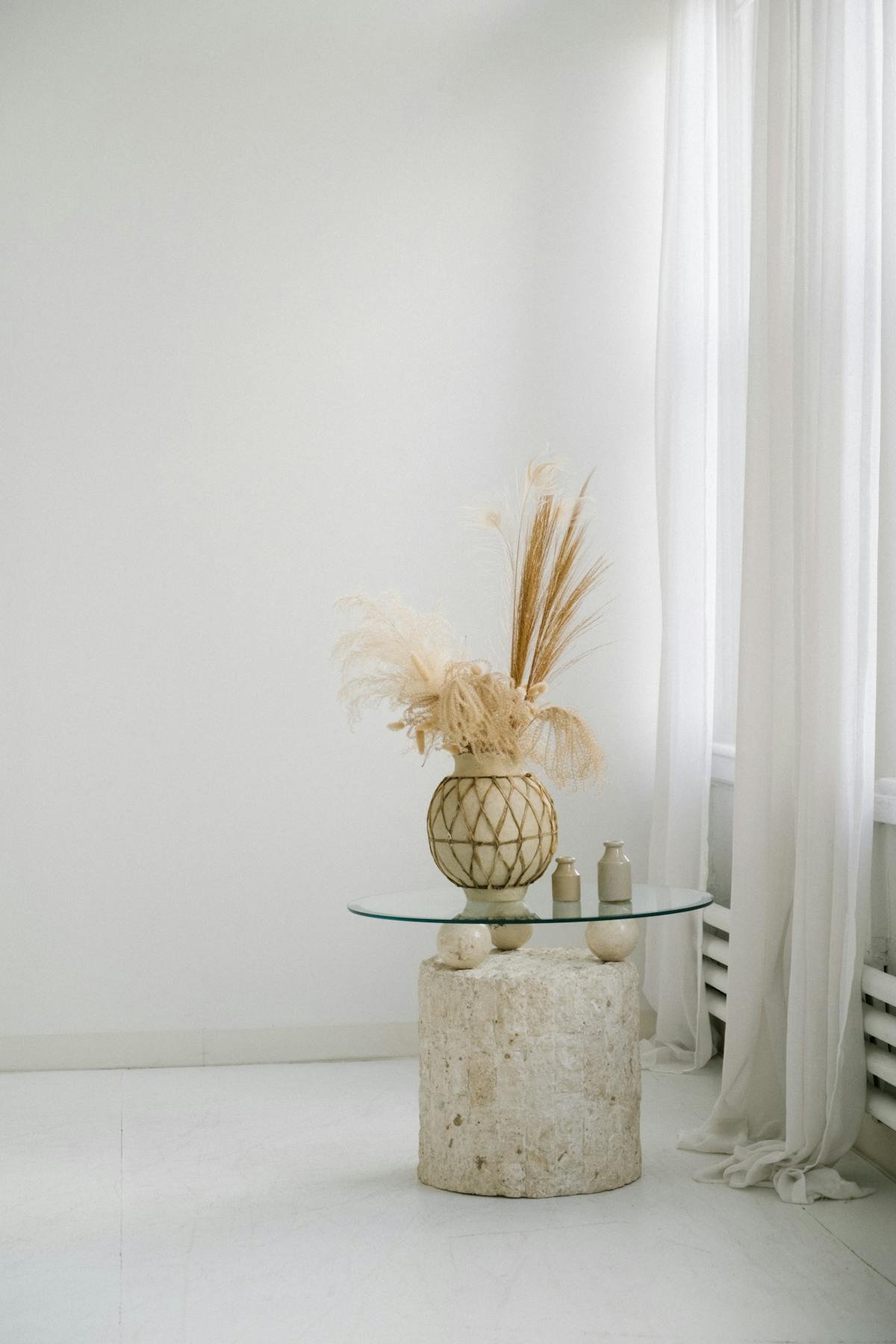 Minimalist interior with a stone pedestal, glass top table, and decorative vases with dried plants, all in neutral tones.