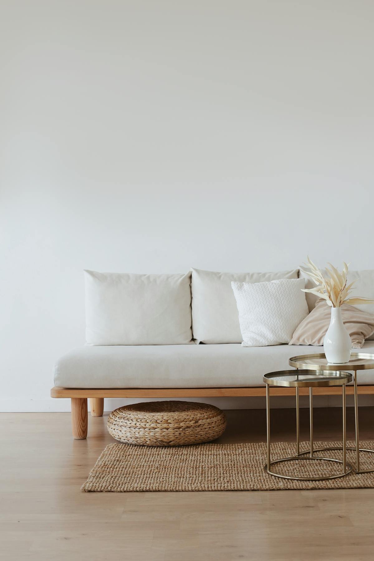 Minimalist living room with a beige sofa, white and beige pillows, a woven round stool under the sofa, and a set of gold side tables with a white vase holding dried leaves. Light wood flooring with a natural fiber rug.