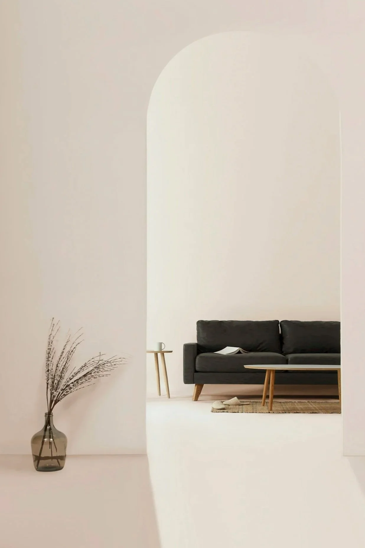 Minimalist living room with a black sofa, wooden coffee table, side table with a mug, and a vase with dried plants on a white wall and floor.
