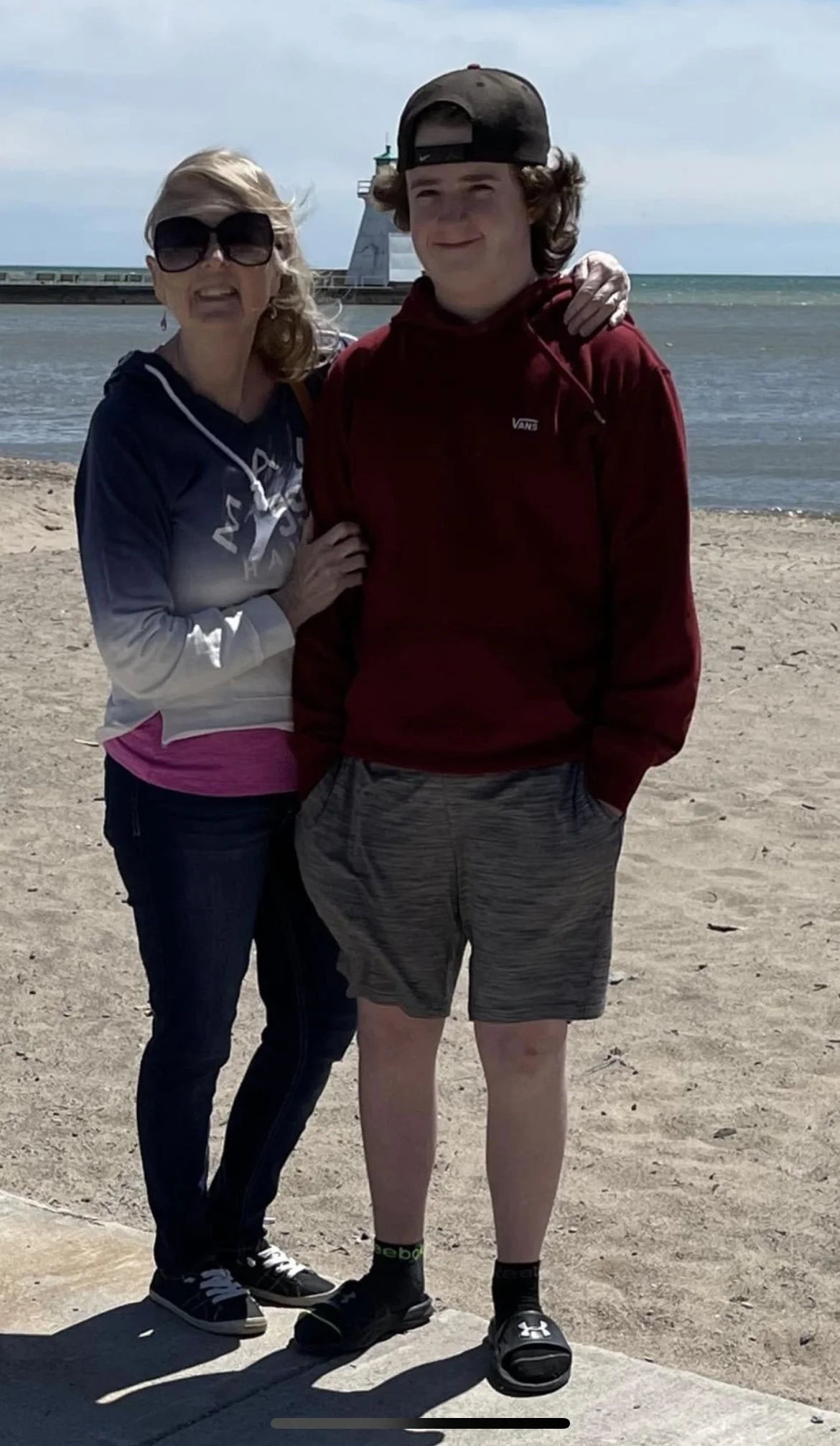 Lucas and Karen Crump standing on a sandy beach with the ocean and a lighthouse in the background. The woman is wearing sunglasses, a hoodie, and jeans, while the boy is wearing a backward cap, a hoodie, shorts, and slides.
