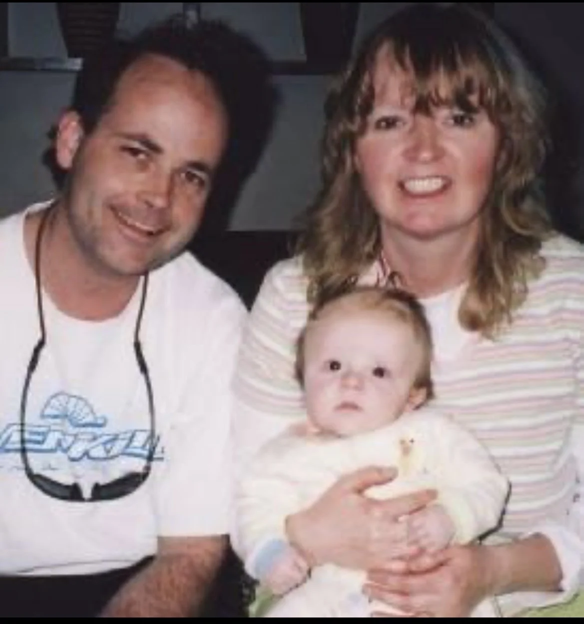 Family photo sitting together and smiling, with the woman holding the baby. The woman has shoulder-length brown hair and is wearing a striped shirt, the man has short dark hair and is wearing glasses around his neck and a white shirt, and the baby ha