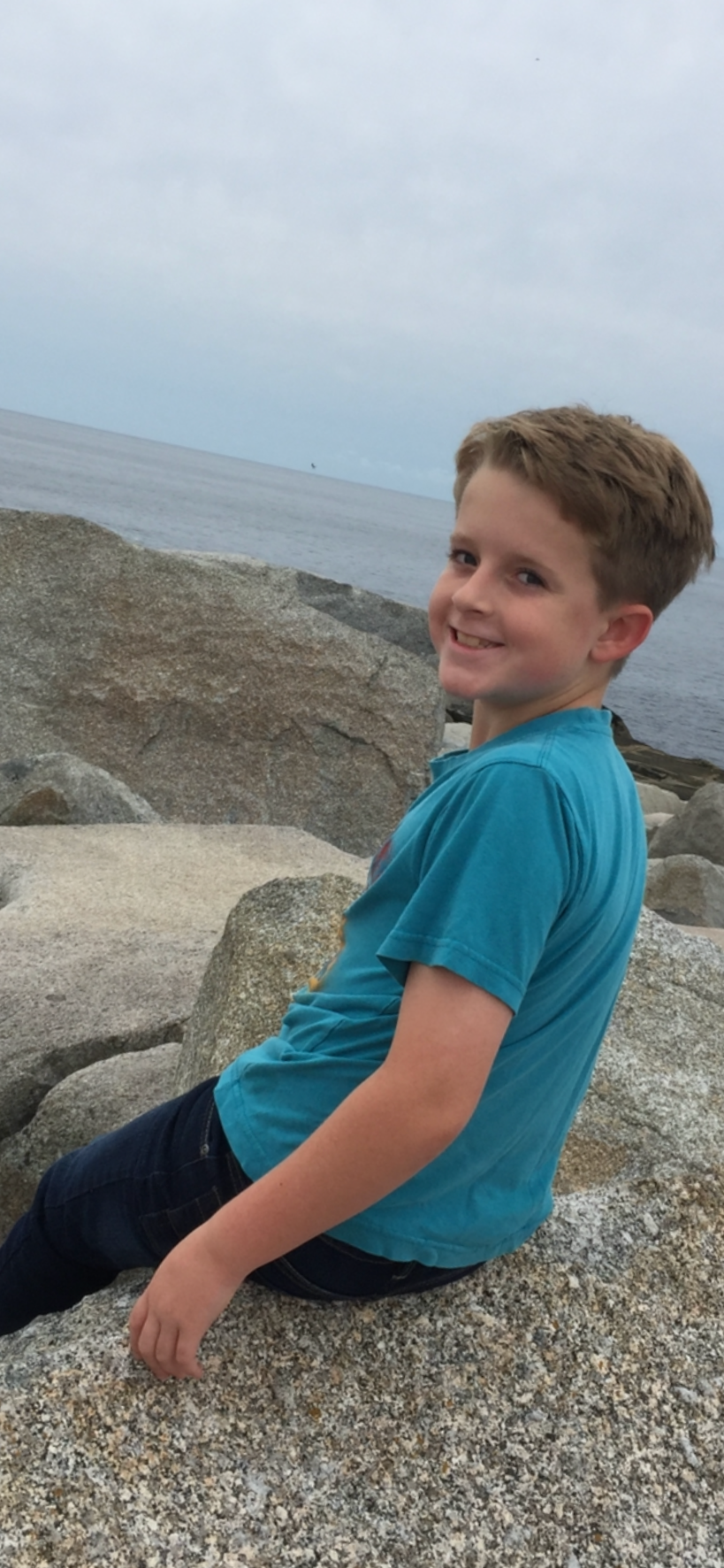 Lucas sitting on rocks by the ocean, smiling at the camera, with a cloudy sky above.