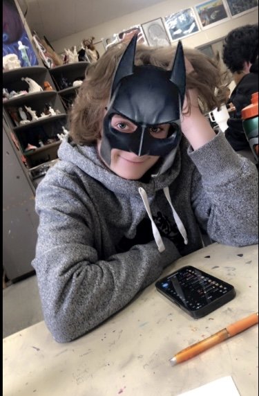 Lucas wearing a Batman mask sitting at a table with a smartphone and a pen, surrounded by shelves filled with various figures and collectibles.