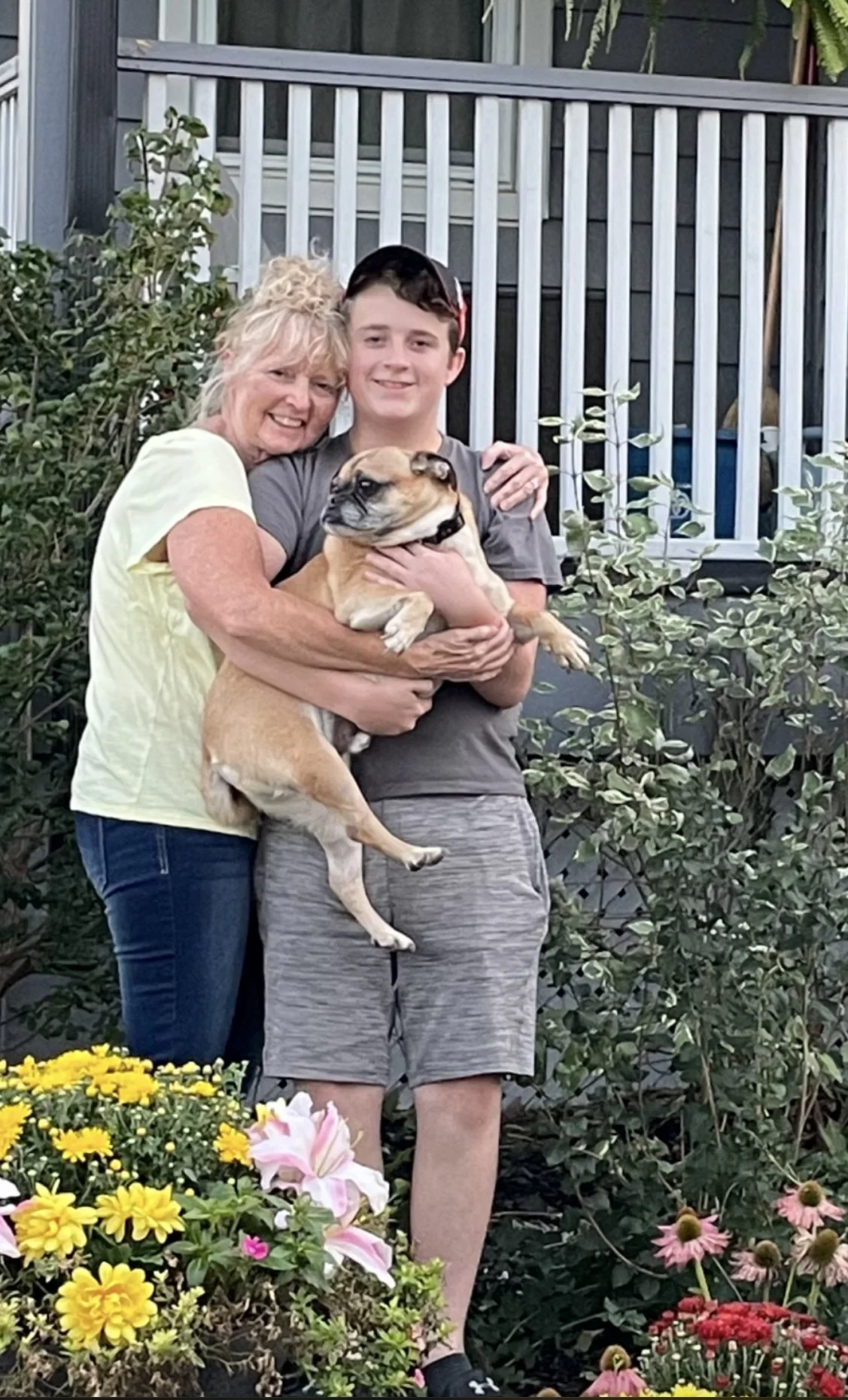 Karen and Lucas holding a bulldog, standing outdoors in a garden with colorful flowers and green bushes, with a house with a porch and white railing in the background.