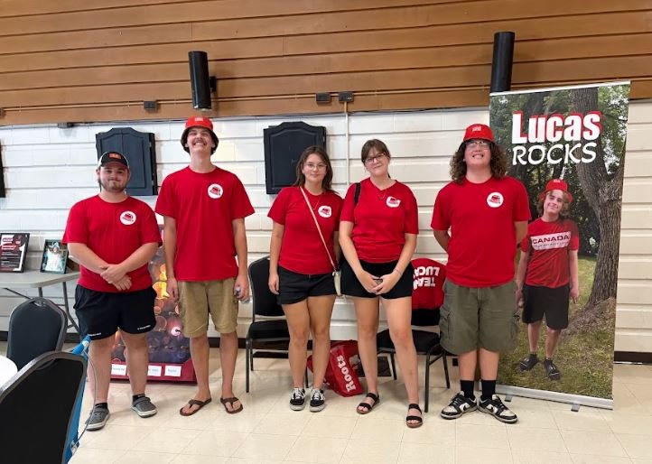 Group of five people standing indoors, wearing red T-shirts and hats, with a banner that says "Lucas Rocks" and features Lucas in a red shirt that says "Canada."