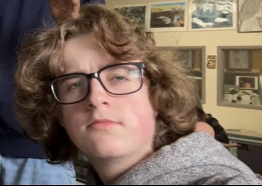 Lucas with curly hair wearing glasses and a grey hoodie, looking directly at the camera with a neutral expression.
