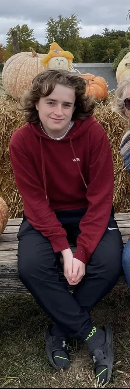 Lucas sitting on a wooden bench at a pumpkin patch, wearing a red hoodie and black pants, with pumpkins and hay bales behind him.