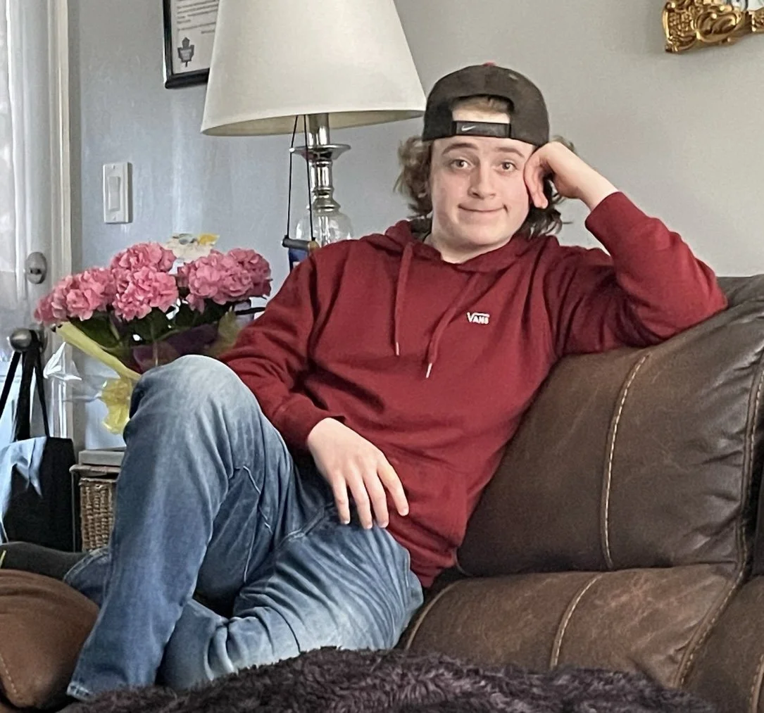 A young man with curly hair wearing a gray baseball cap backwards, a red hoodie, and blue jeans, sitting on a brown leather couch with one arm resting on the armrest and his head leaning on his hand, smiling at the camera in a living room with pink f