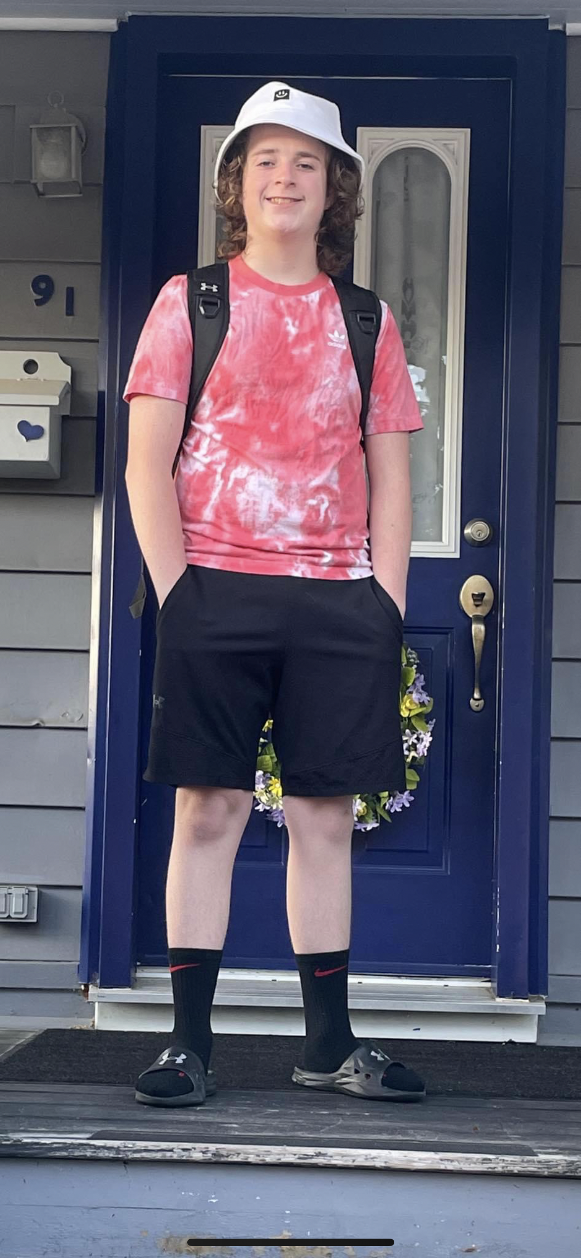 Lucas with curly hair stands in front of a blue door with a smile, hands in his shorts pockets. He wears a white bucket hat, a red tie-dye T-shirt, black shorts, black Nike socks, black sandals, and a black backpack.