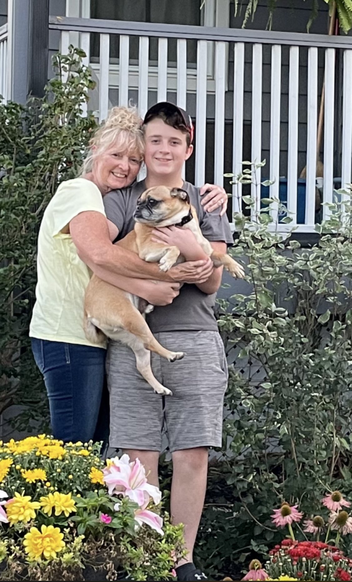 Karen and a Lucas are standing outside on a patio or garden area, holding a small tan French Bulldog. Behind them is a white porch railing, green leafy plants, and colourful flowers in the foreground.