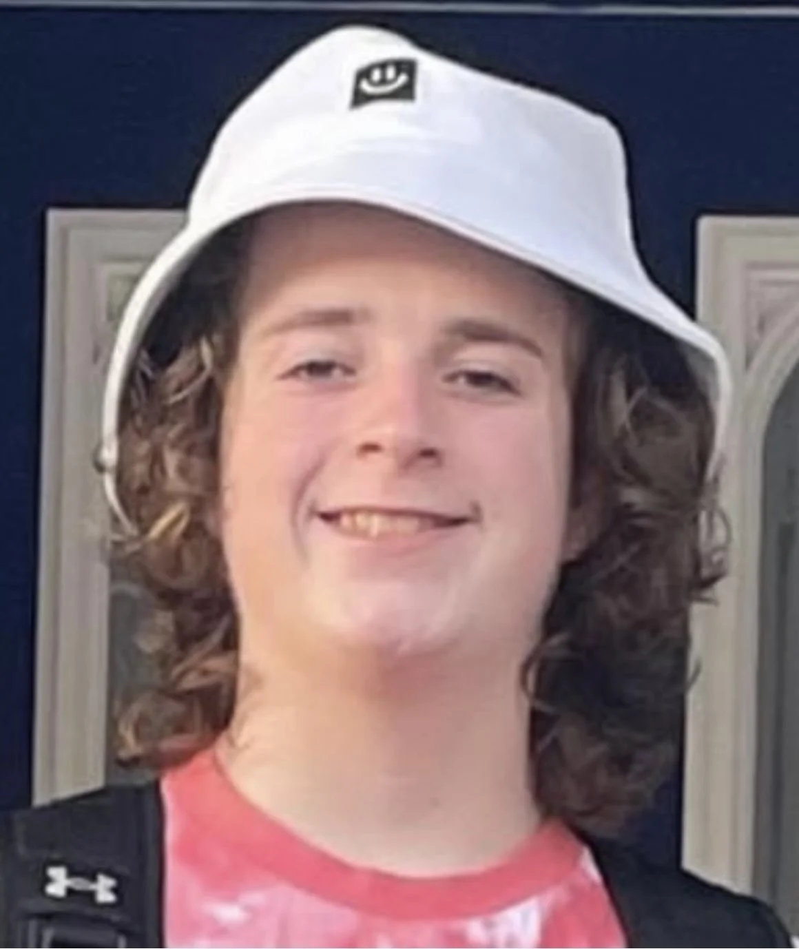 Lucas Crump, wearing a white bucket hat, pink tie-dye shirt, and black backpack, smiling in front of a dark door.