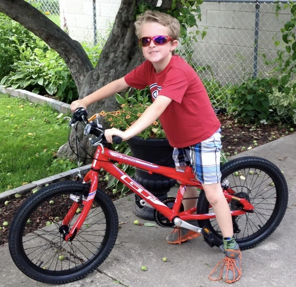 Young Lucas wearing sunglasses, a red t-shirt, and plaid shorts, standing next to a red bicycle on a sidewalk. There are plants, a tree, and a fence in the background.