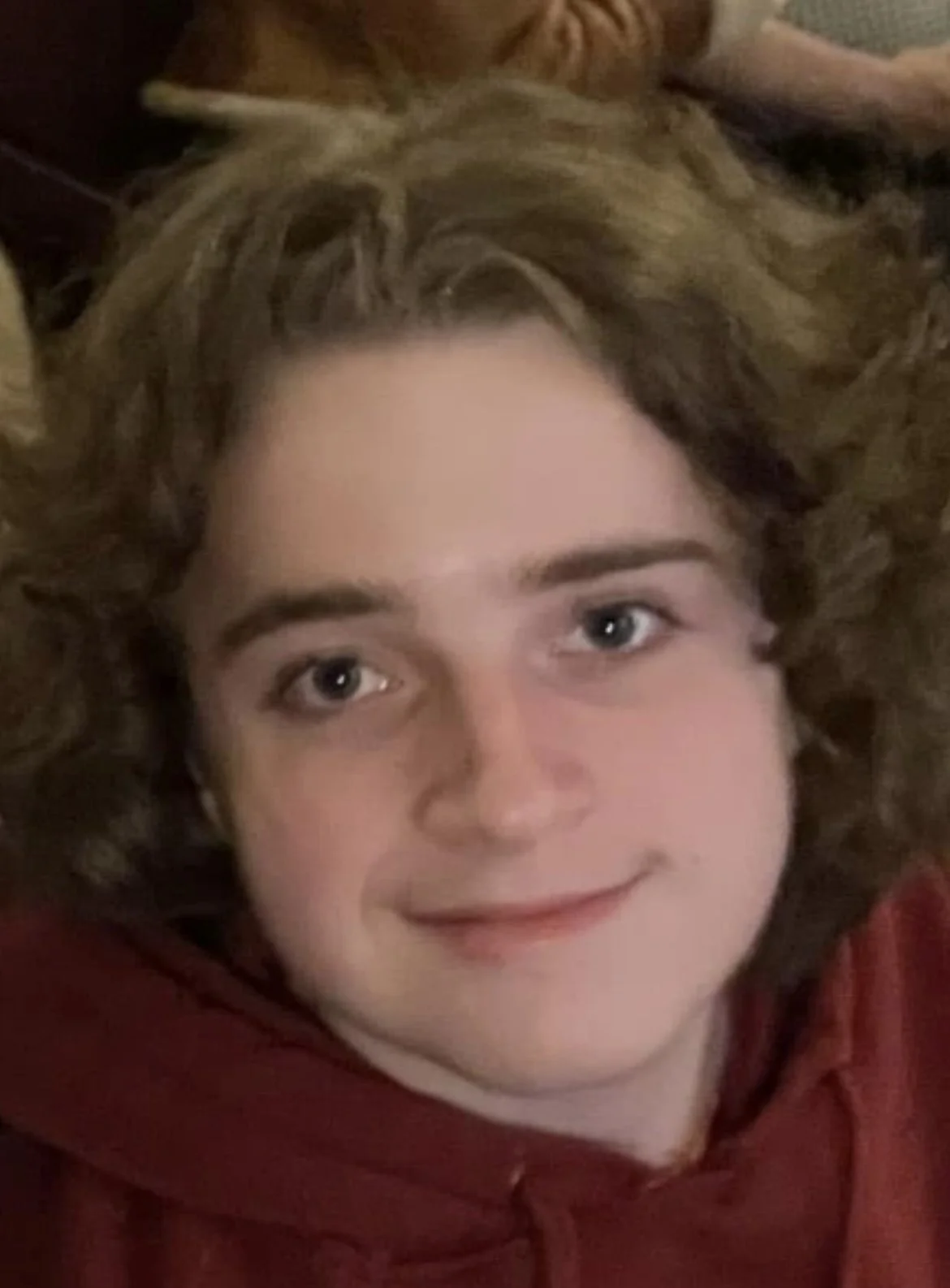 Close-up of Lucas lying down with curly brown hair, wearing a red hoodie, and smiling slightly.