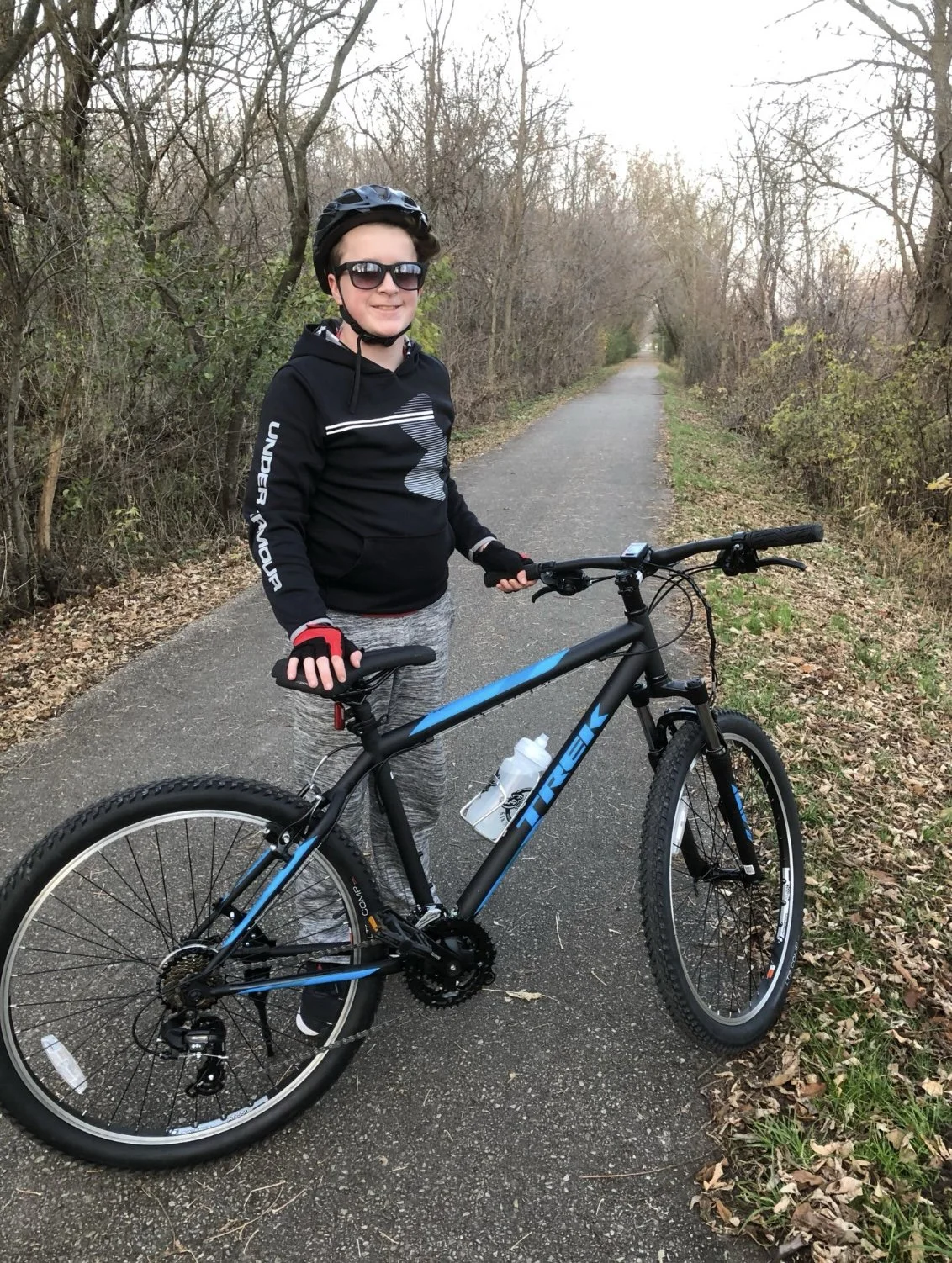 Lucas wearing sunglasses, a black helmet, and black and gray athletic clothing stands on a paved trail with a black and blue mountain bike, holding the handlebars, in a wooded area with trees and fallen leaves.