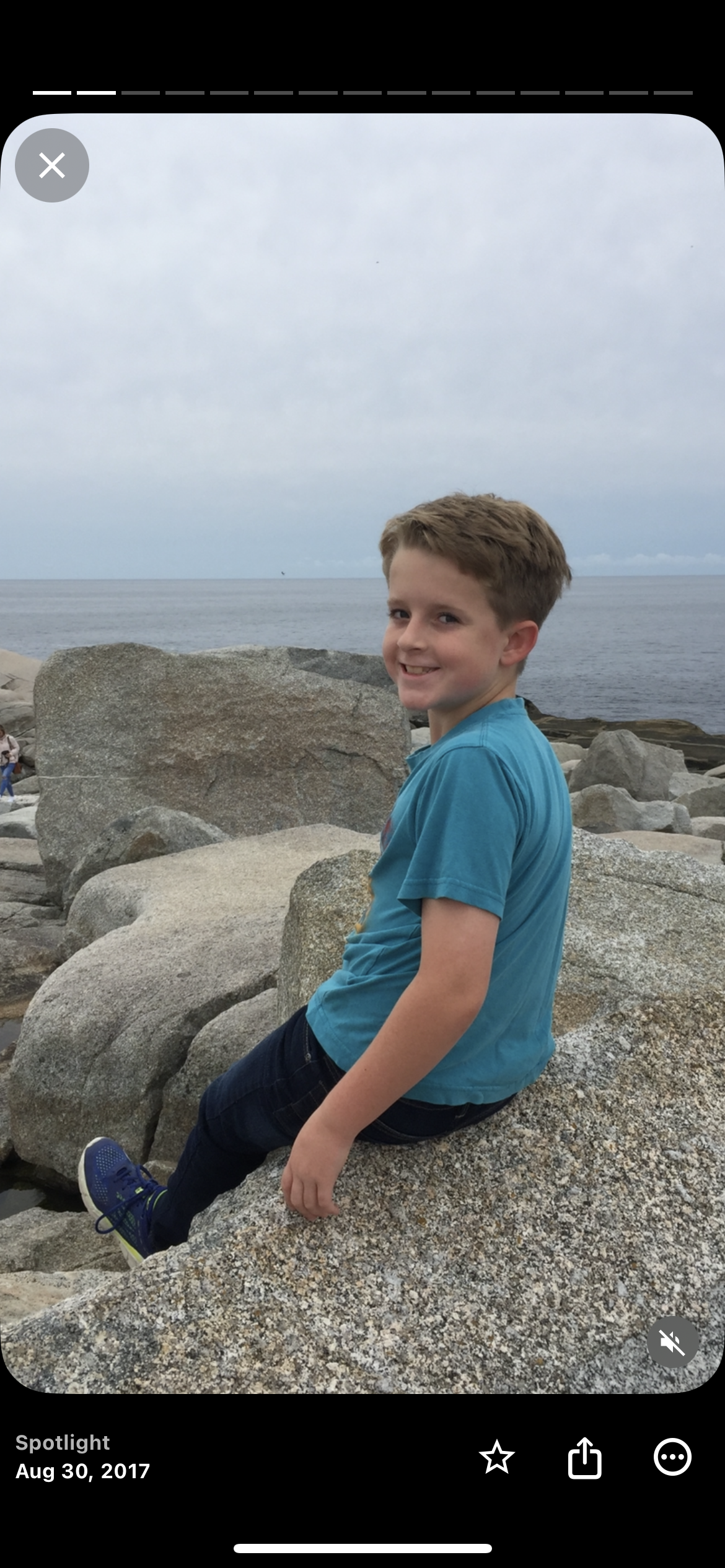 Lucas, wearing a blue t-shirt, black pants, and blue shoes, sitting on large rocks near the shoreline with the ocean and cloudy sky in the background.