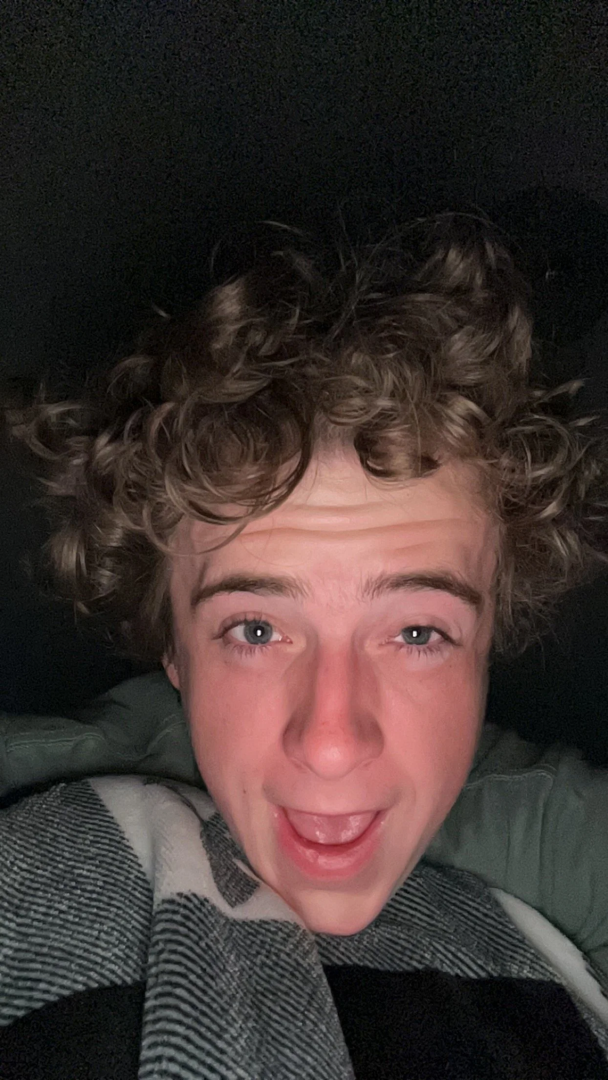 Lucas with curly hair, smiling, lying on a dark surface, wearing a striped shirt and a green jacket.