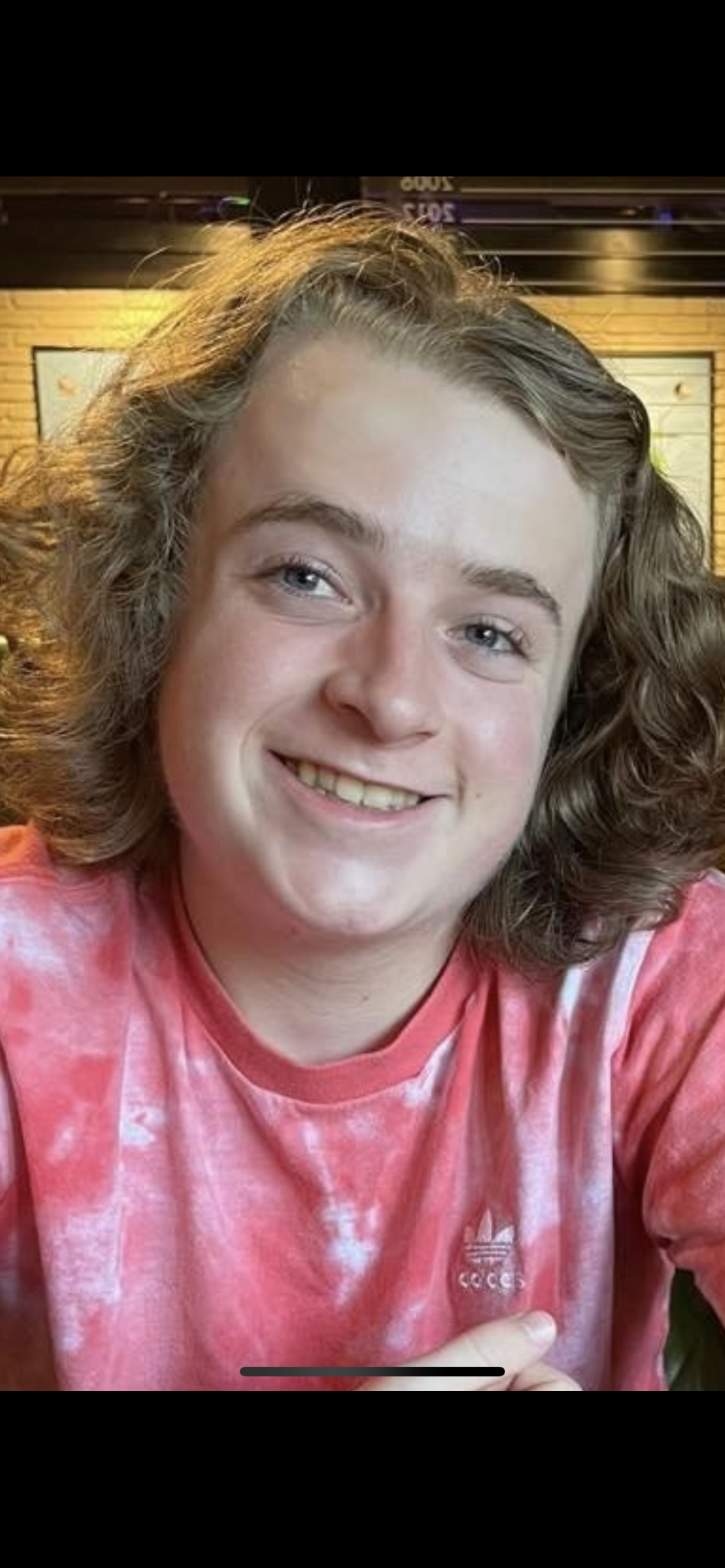 Lucas with shoulder-length curly hair smiling at the camera, wearing a pink tie-dye Adidas shirt, in a restaurant or cafe setting.