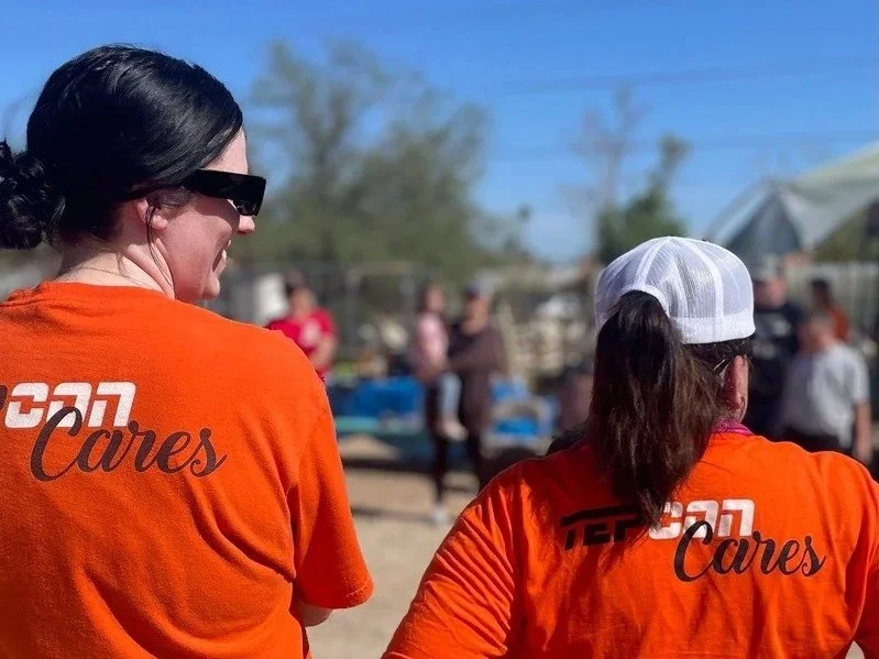 Two women wearing orange shirts with 'TEPCON Cares' printed on the back, outdoors on a sunny day, with a crowd of people in the background.