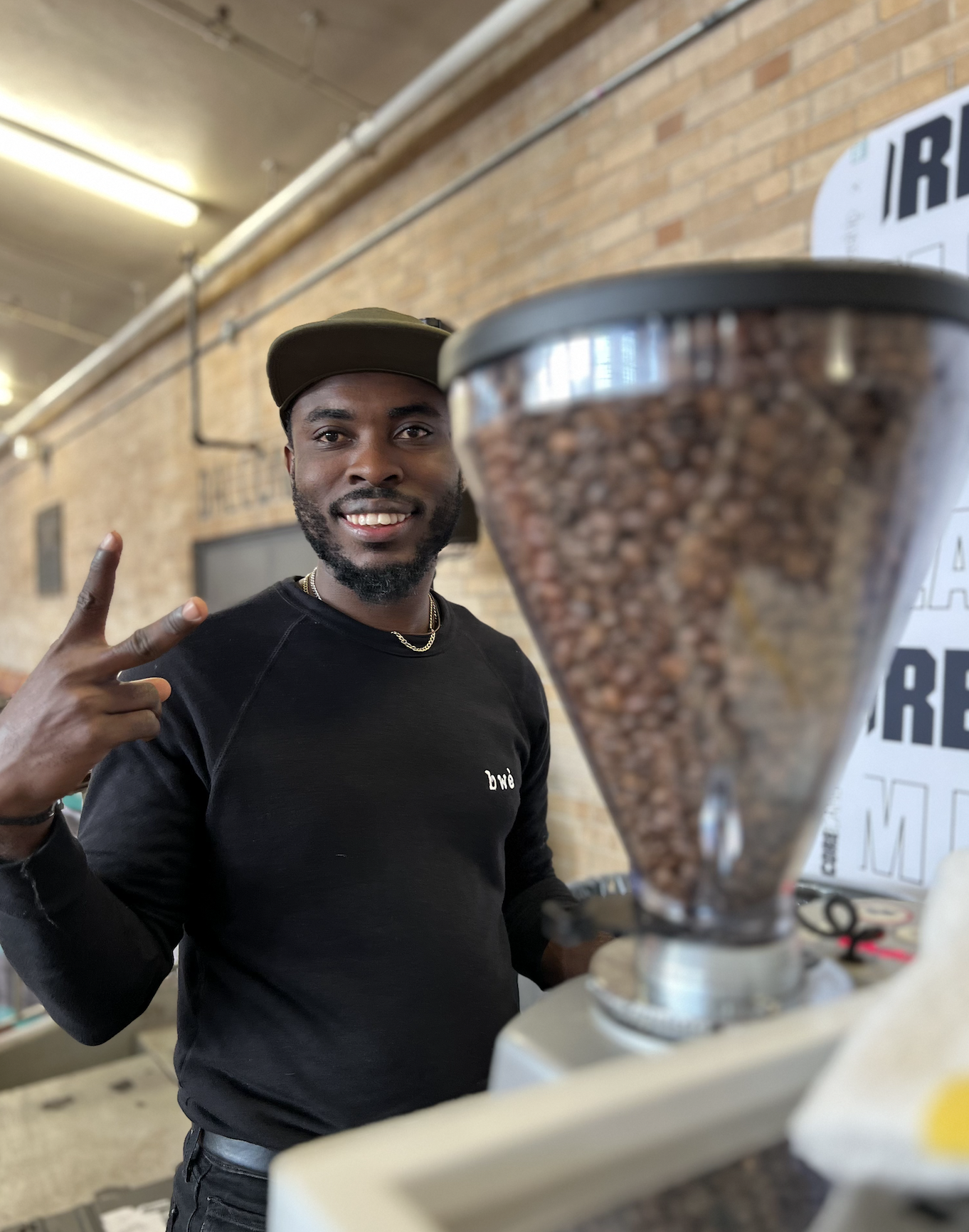 Man smiling and making a peace sign next to a coffee grinder in a cafe with a brick wall background.