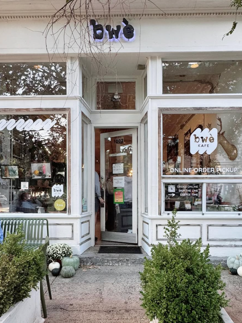 White storefront with large glass windows advertising BWE KAFE, featuring signs for online order pickup and skip the line coffee. The entrance has a partially open door with a person inside. Outside, there are green plants and pumpkins decorated on either side of the entrance, along with a green bench.