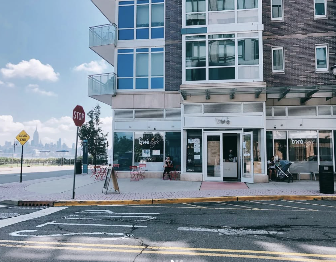 A city street scene featuring a café called 'bwe' with outdoor seating, a red stop sign, a yellow dead end sign, and a skyline of skyscrapers in the background under a partly cloudy sky.