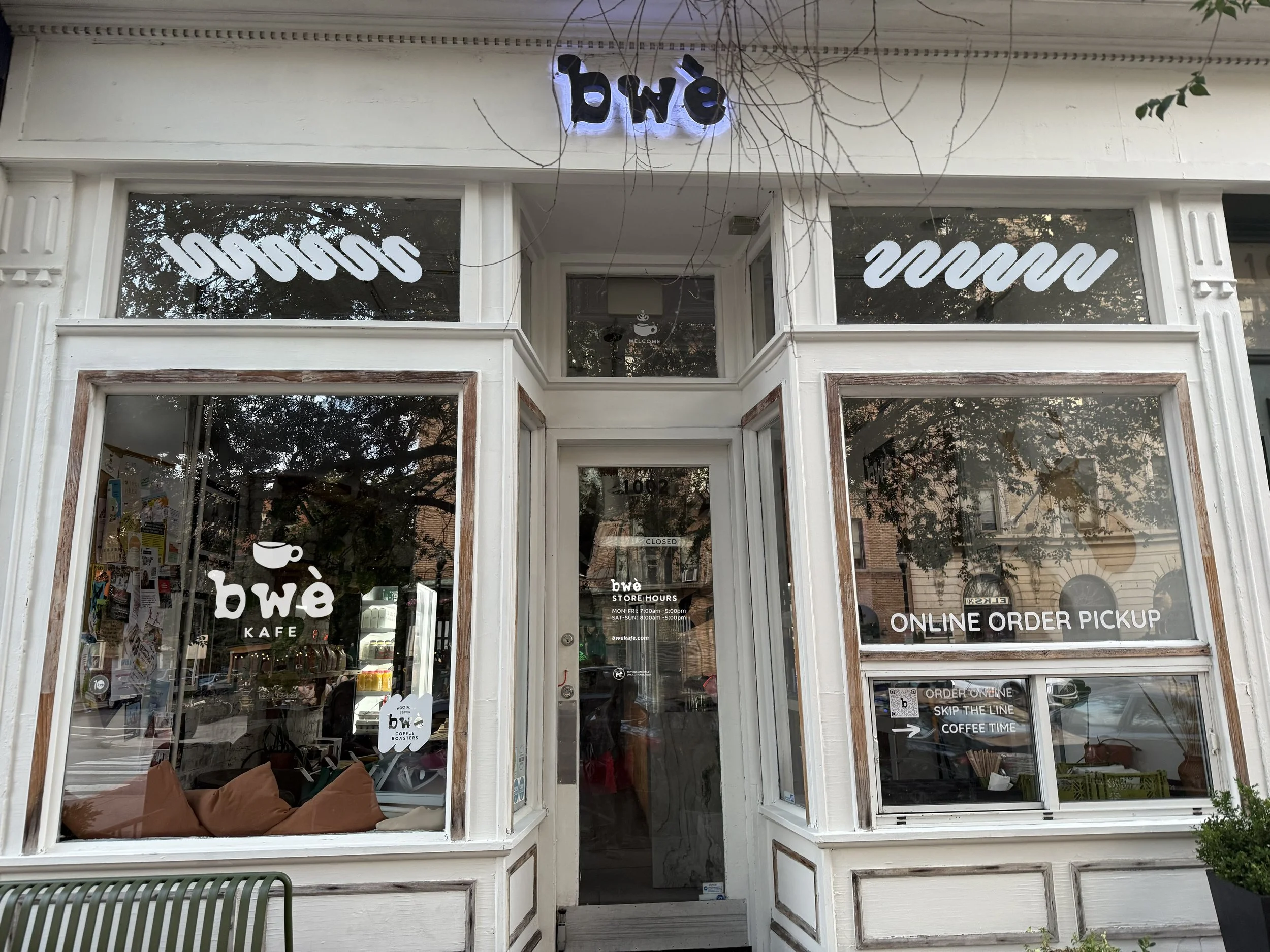 Front view of a coffee shop with large windows displaying the store's name 'bwe,' window signs indicating 'ONLINE ORDER PICKUP' and 'ORDER ONLINE SKIP THE LINE COFFEE TIME,' and the store hours posted on the door.