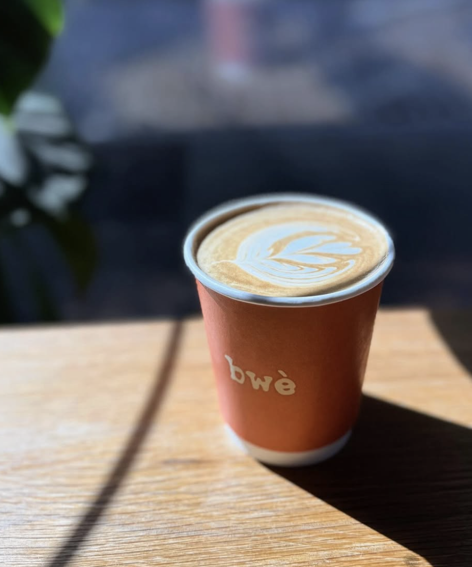 A paper cup of coffee with latte art sitting on a wooden table, sunlight casting a shadow.