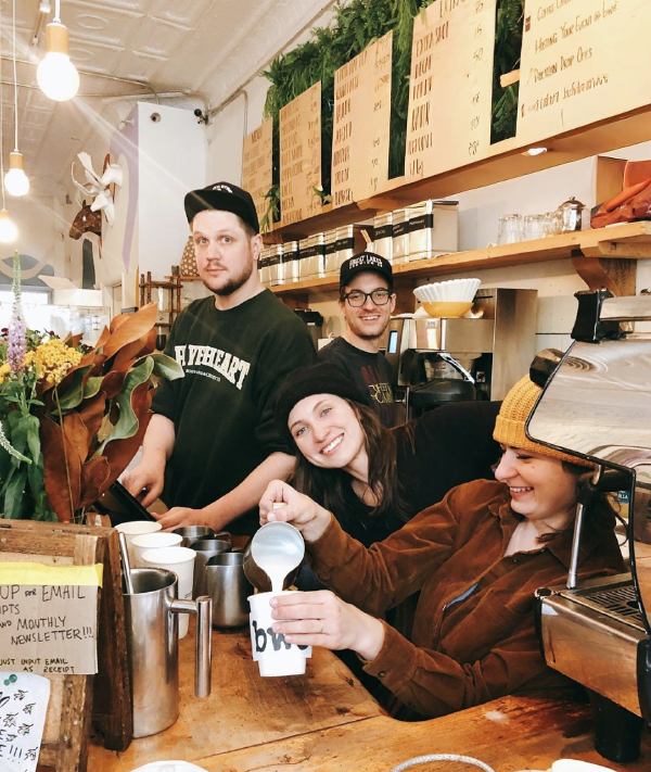 Four baristas behind a coffee counter, smiling and posing for a photo inside a cozy café with wooden shelves, plants, and a menu board.