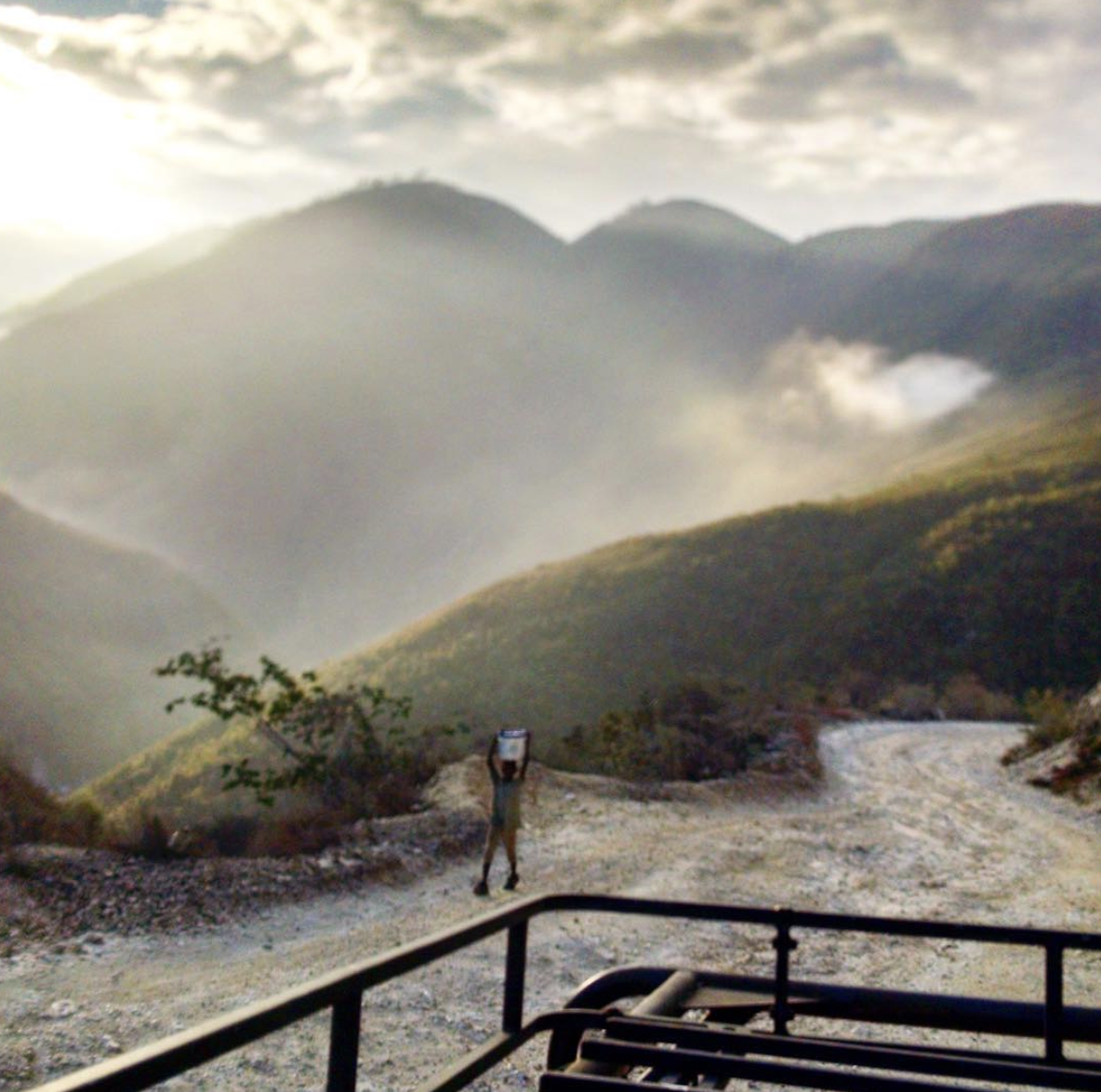 A person standing on a dirt road in a mountainous area, holding a camera or phone to take a photo with mountains and cloudy sky in the background.