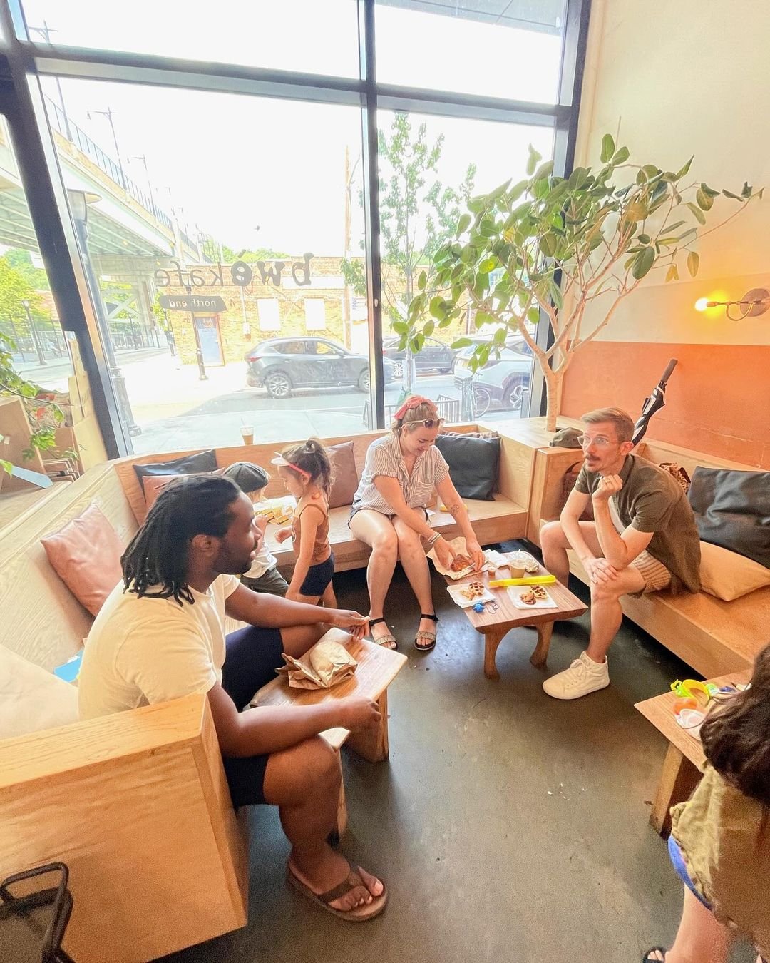A group of people, including two children, sitting in a cozy cafe with large windows, wood furniture, and a potted plant inside. They are eating food and talking.