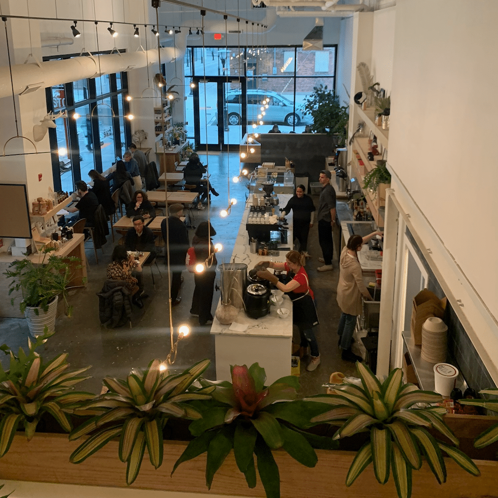 Interior of a busy coffee shop with customers seated at tables, staff preparing drinks behind the counter, large windows letting in natural light, and decorative plants near the front.