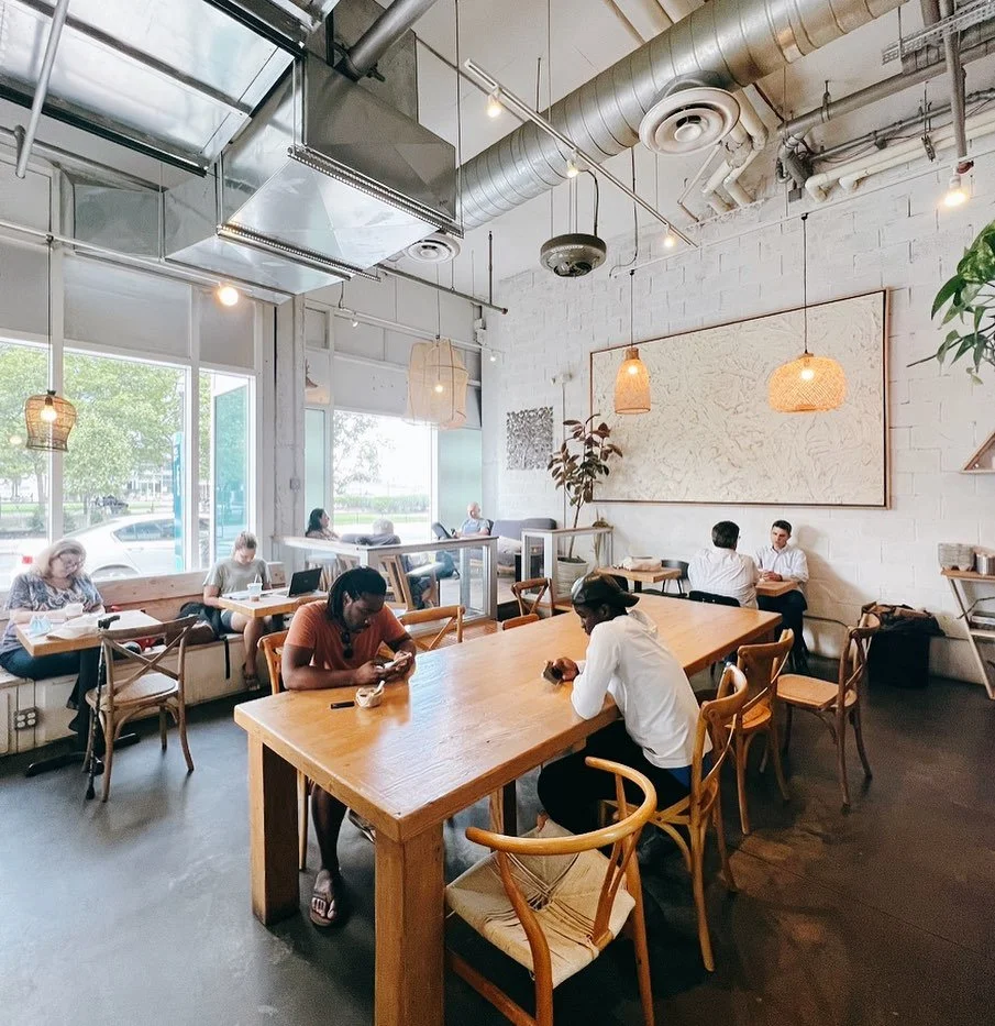 Interior of a modern cafe with several customers seated at tables, large windows letting in natural light, and minimalist decor including hanging light fixtures and a large textured artwork on the white brick wall.