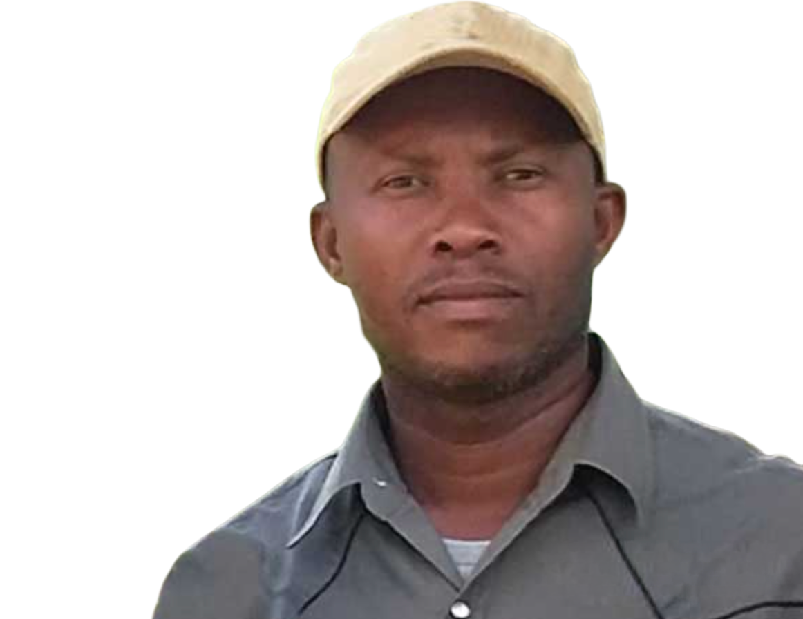 A man wearing a tan cap and navy blue shirt standing outdoors with a serious expression on his face.