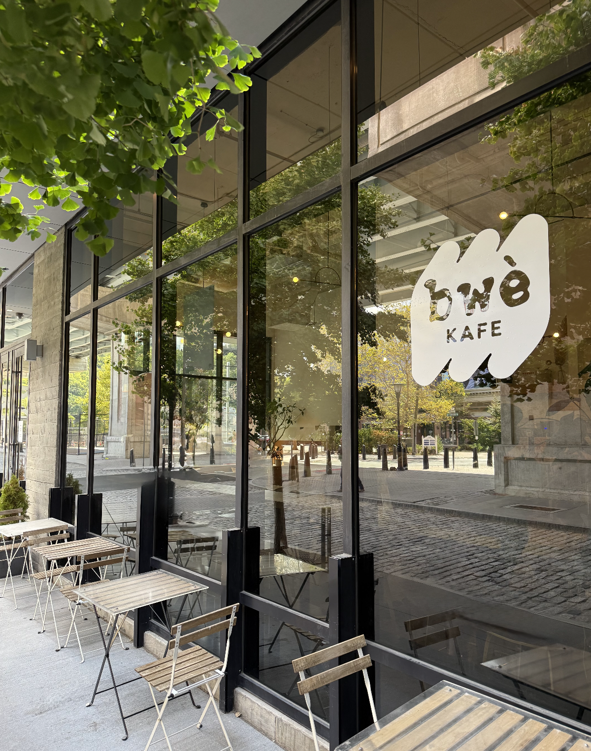 A cafe with large glass windows, outside seating with wooden tables and chairs, and a sign that reads 'bwé KAFE' on the window. There are trees and a cobblestone street visible through the glass.