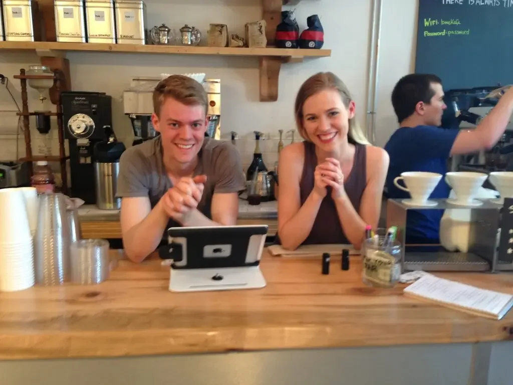 A man and woman smiling while sitting behind a coffee shop counter, with coffee cups and brewing equipment visible.