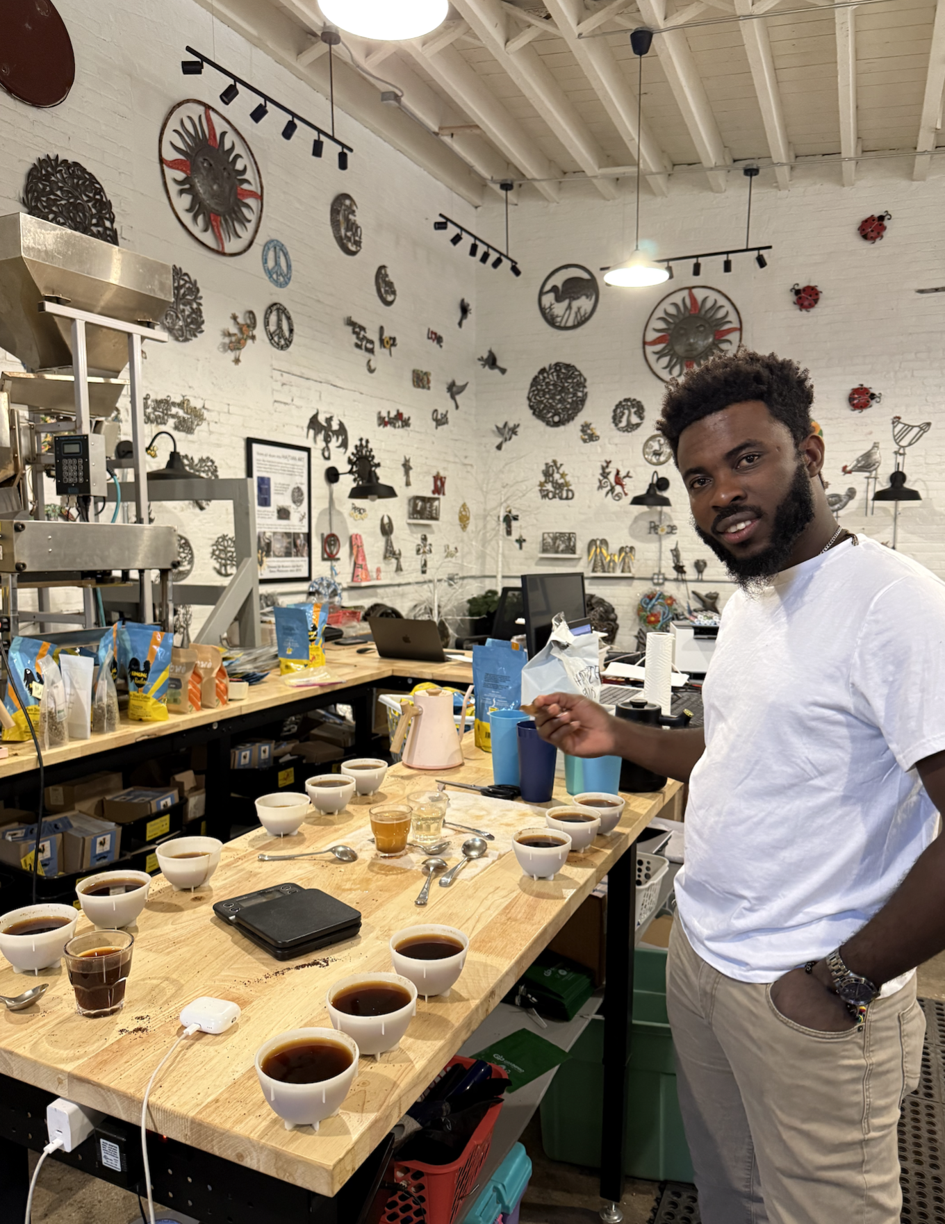 A man in a white t-shirt standing at a wooden table with multiple cups of coffee, spoons, and a scale, in a cafe with wall art, computers, and coffee supplies in the background.