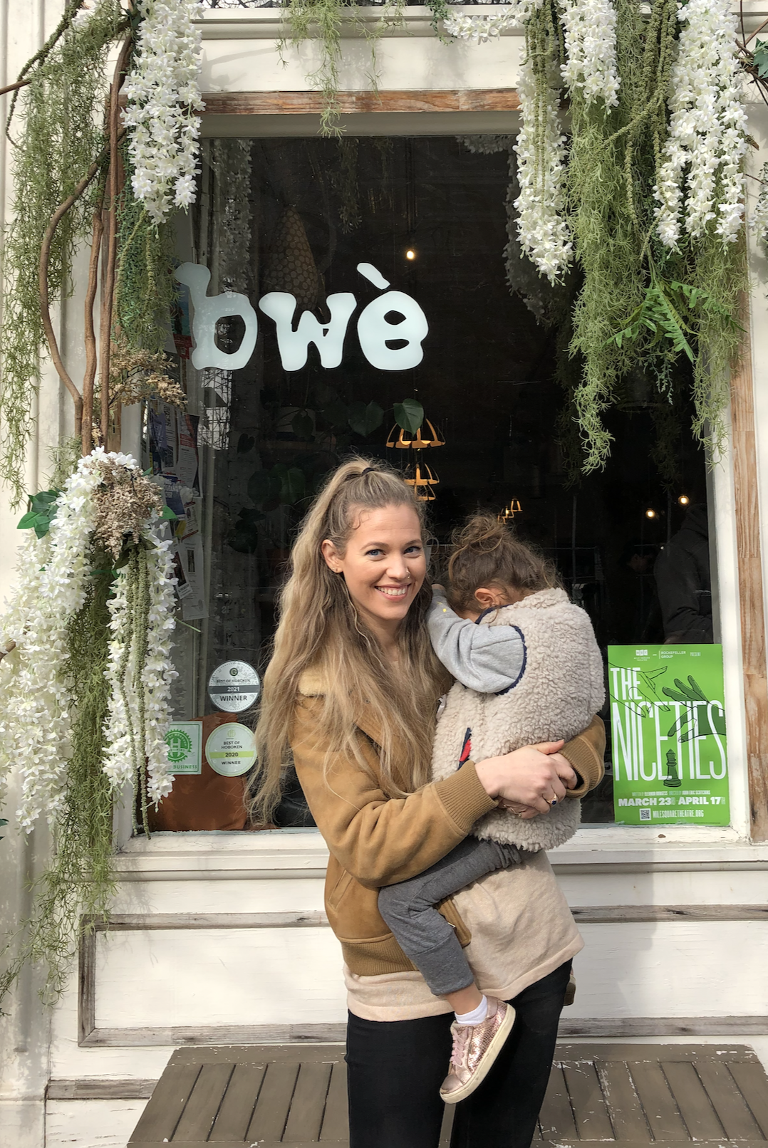 Smiling woman holding a young girl outside a flower shop with white flowers and green foliage around the window, and a green poster inside advertising 'The Nites' event from March 23 to April 17.