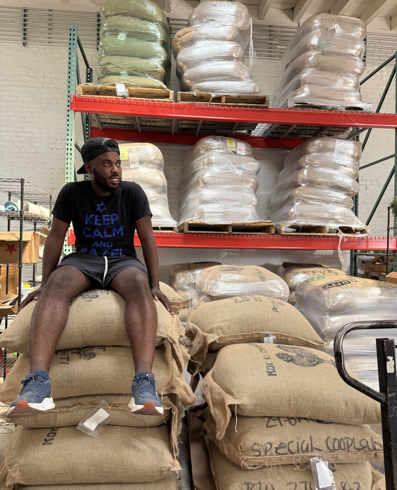 A young man sitting on a large sack of coffee beans in a warehouse surrounded by stacked burlap and plastic-wrapped sacks of coffee.