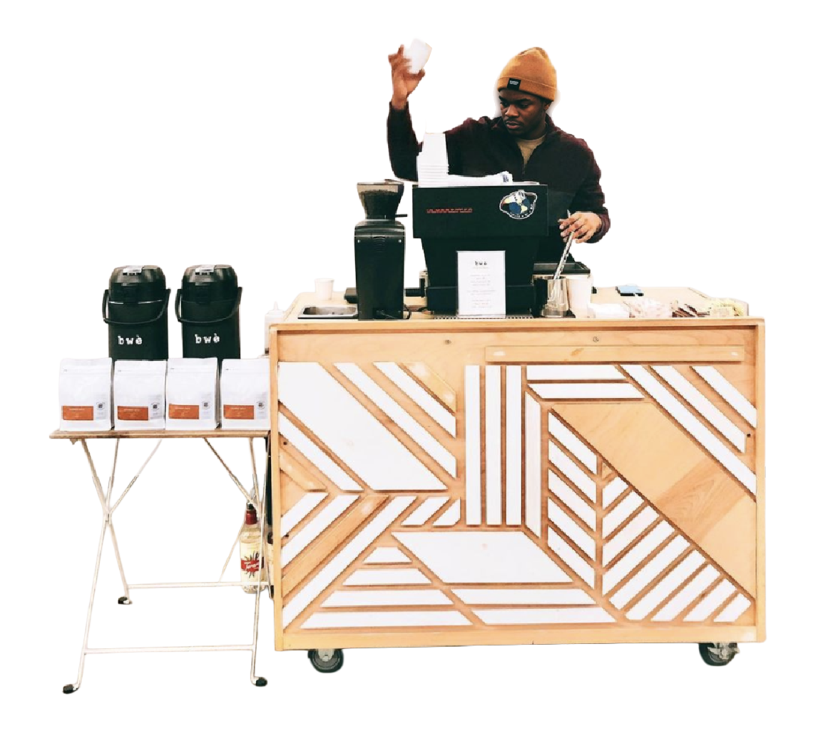 Barista preparing coffee at a modern wooden coffee stand with geometric design, two black coffee dispensers, coffee bags, and a small table with additional coffee supplies.