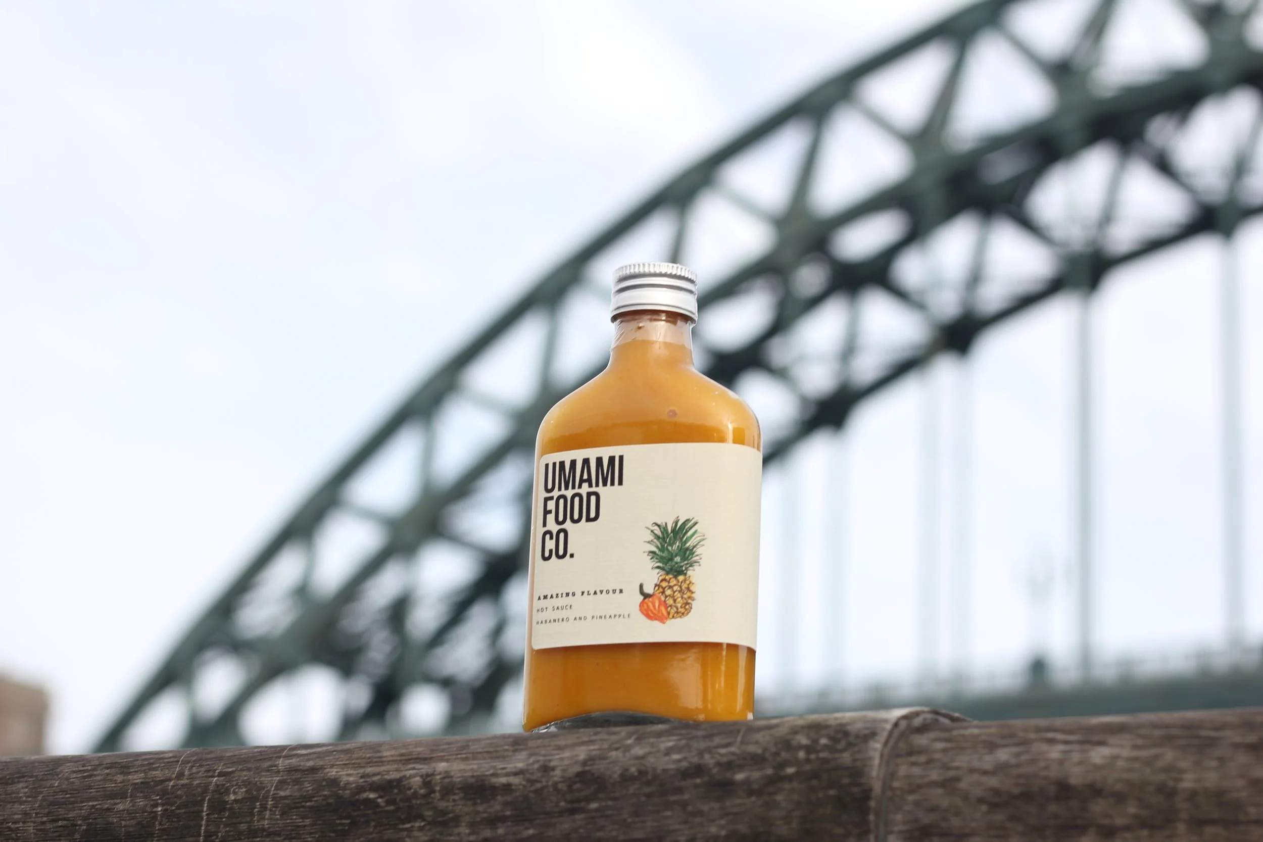 Bottle of orange sauce labeled "Umami Food Co." with a pineapple and chili illustration, placed on a wooden surface with a bridge in the background.