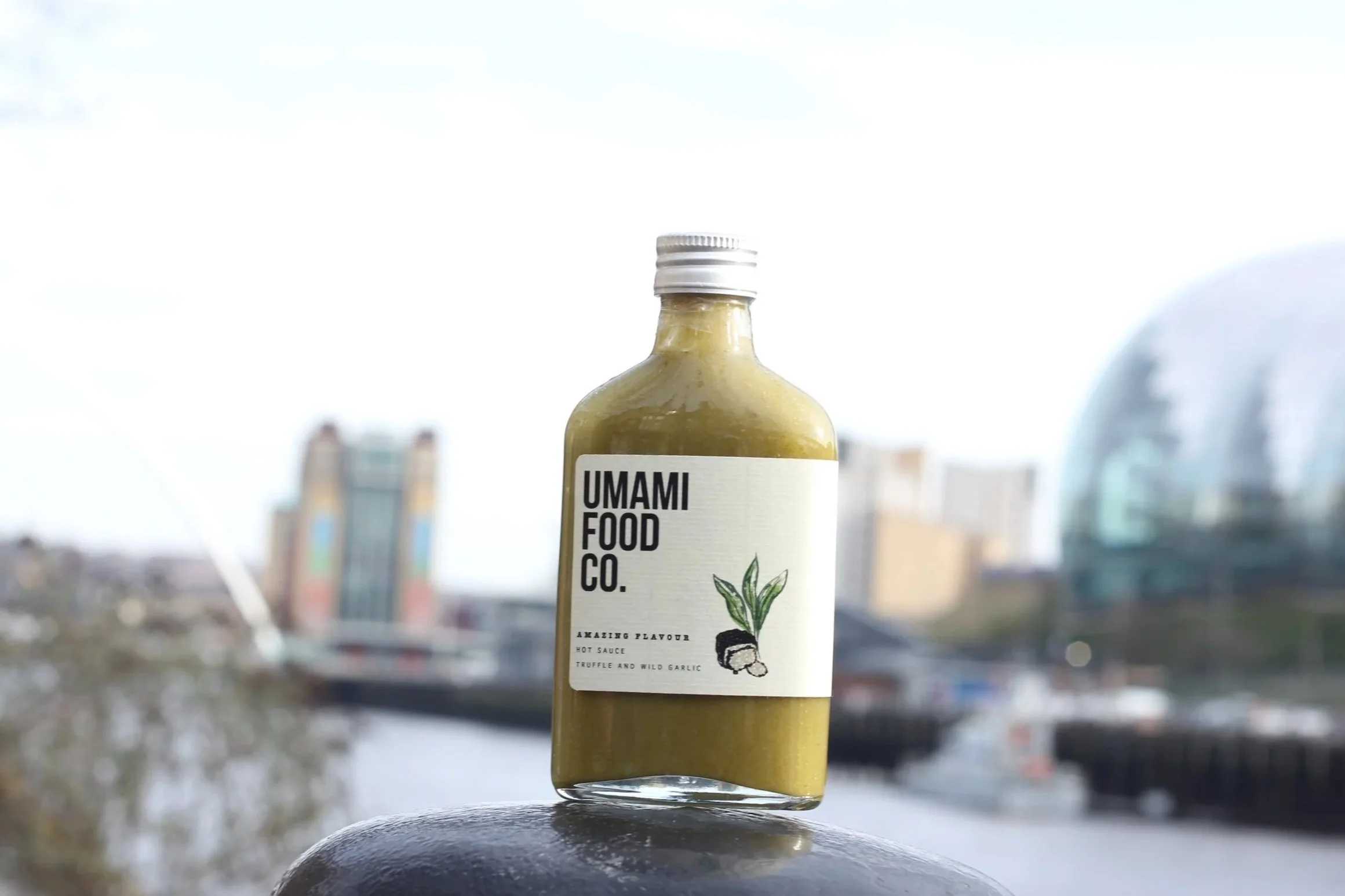 A bottle of Umami Food Co. condiment with a label featuring a leaf and garlic illustration, set outdoors with an urban skyline and water in the background.