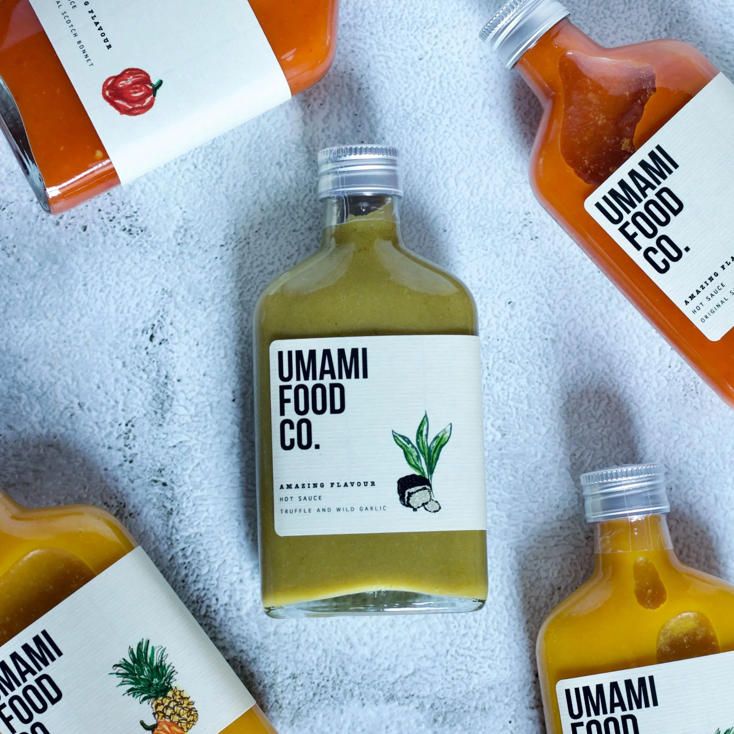 Four bottles of Umami Food Co. hot sauce with various flavors, one in the center showing a greenish sauce with a label featuring a green leaf and a black and white illustration, the others around it showing different sauces with labels and illustrations of a red pepper, a pineapple, and a yellow substance.