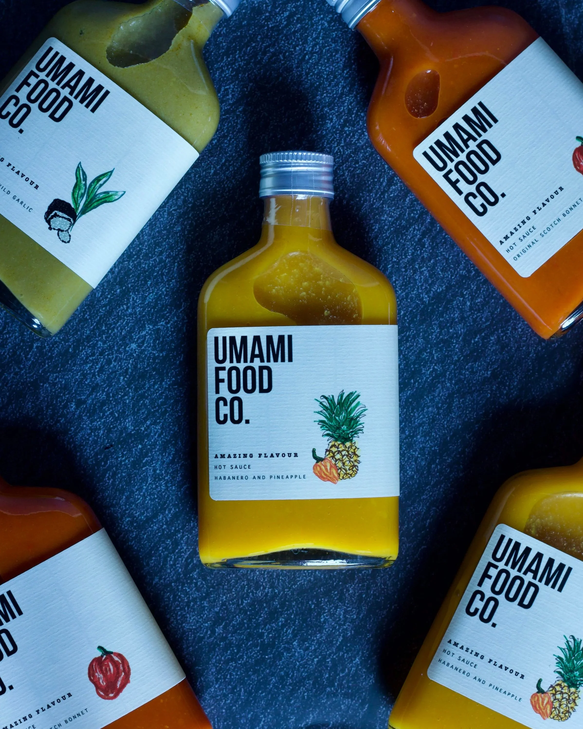 Bottles of hot sauce from Umami Food Co. with labels showing different flavors, including habanero and pineapple, on a dark textured surface.