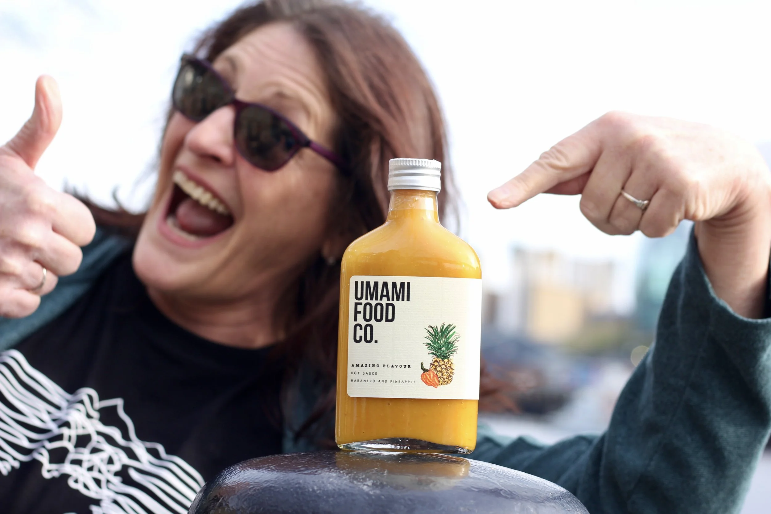 A smiling woman with sunglasses giving a thumbs-up and pointing at a bottle of UMMAMI FOOD CO. hot sauce on a surface outdoors.