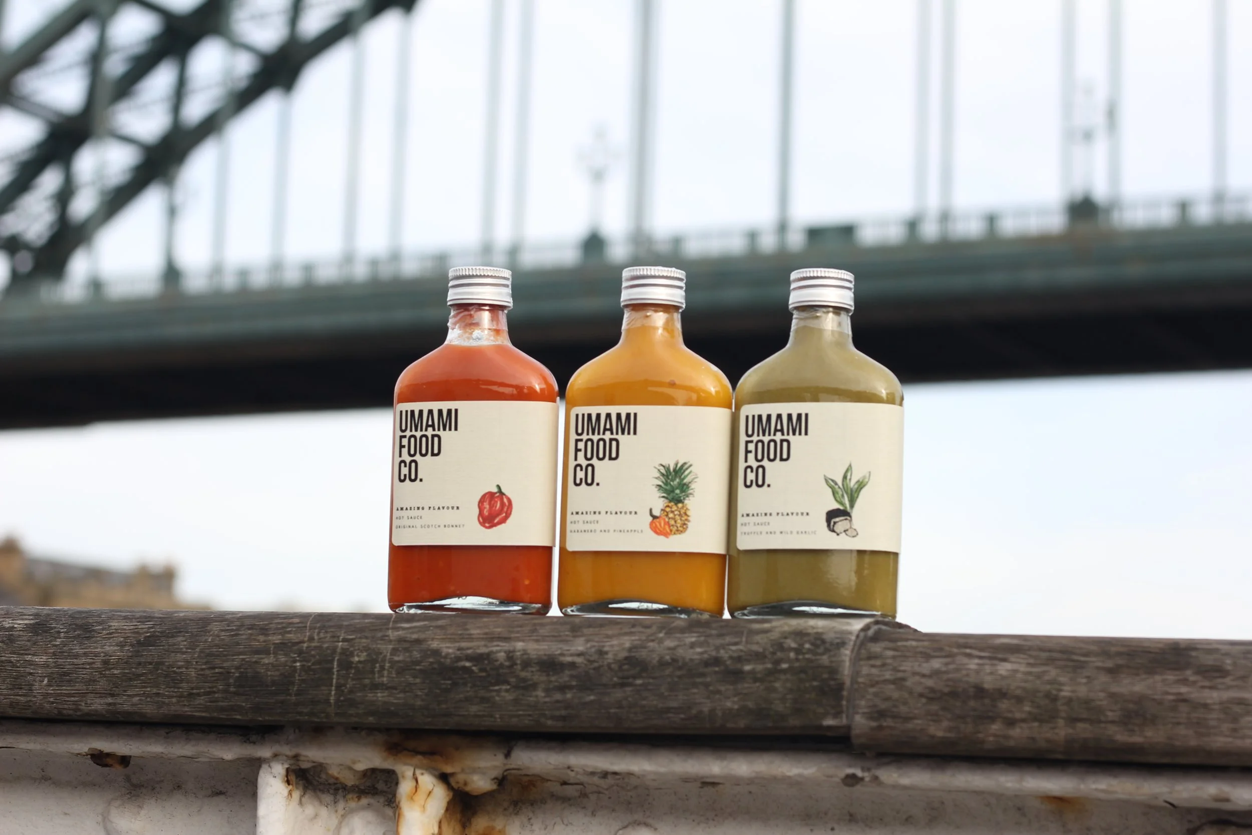 Three bottles of food sauce with labels featuring images of a red pepper, pineapple, and ginger, placed on a wooden surface with a bridge in the background.