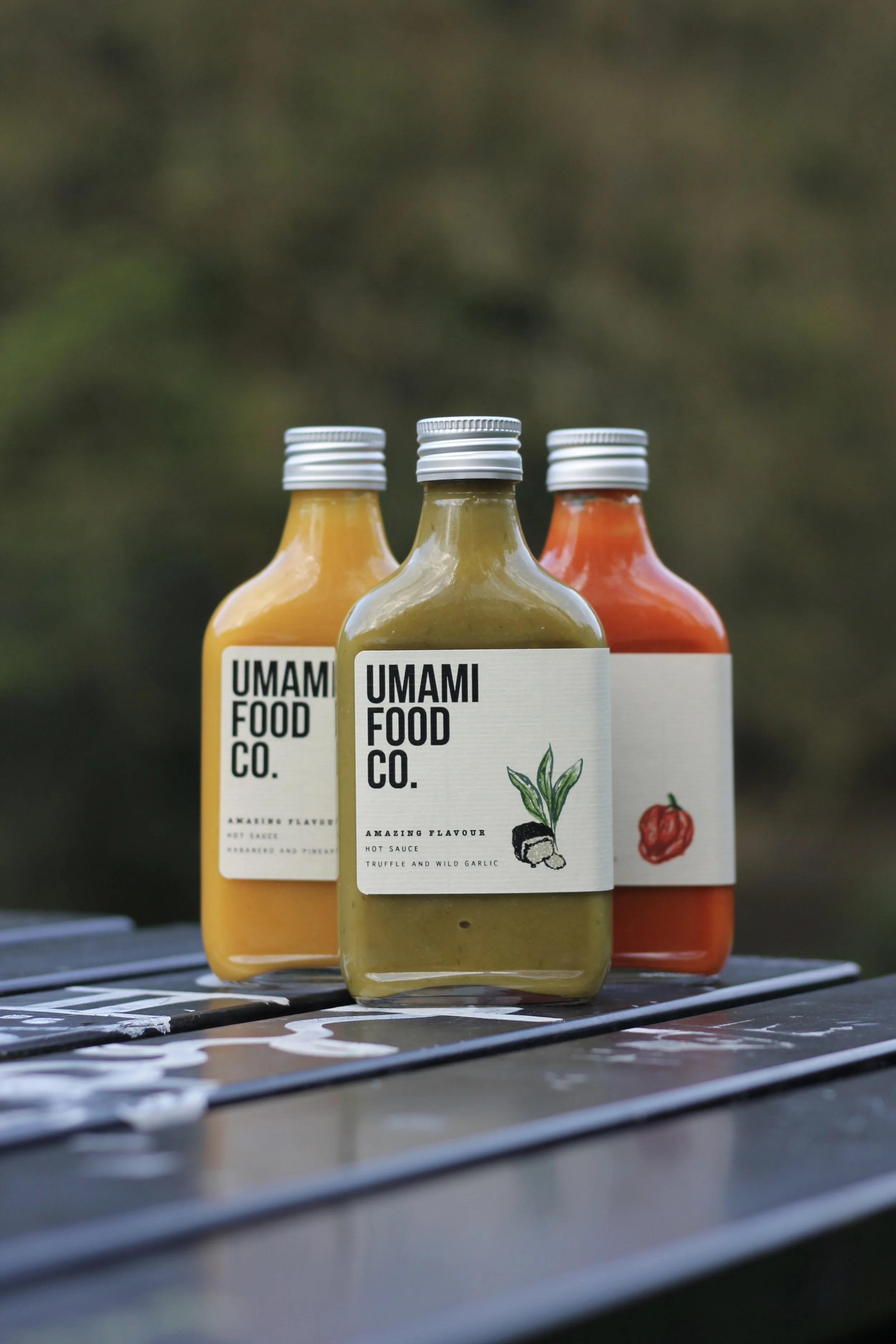 Three bottles of hot sauce from Umami Food Co. are placed on a metal surface outdoors. The bottles are different colors, with the front one being green, the left yellow, and the right red, each labeled with the flavor and ingredients.