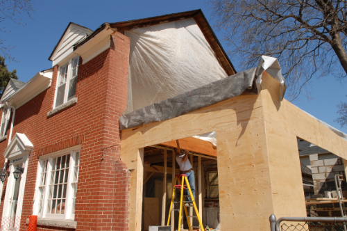 A house is under construction with a brick exterior and new wooden framework being added. A worker is on a ladder working on the structure, and some trees and a clear blue sky are visible in the background.